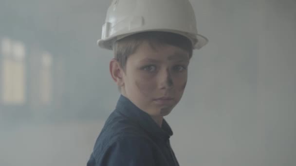 Portret schattig jongen in een beschermende helm draaien en kijken naar de camera op de achtergrond van rook binnenshuis. Concept van brand, ramp, ontvlambaarheid, niet-naleving van veiligheidsvoorschriften. — Stockvideo
