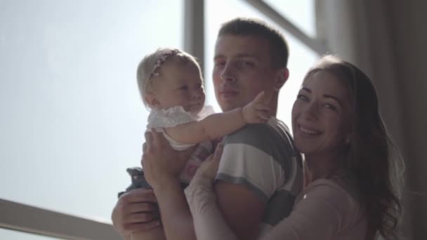 Beau jeune homme tenant une petite fille dans ses bras devant la fenêtre gros plan. Maman est à côté de son mari et de son bébé. Les joies de la paternité. Famille aimante à la maison. Mouvement lent — Video