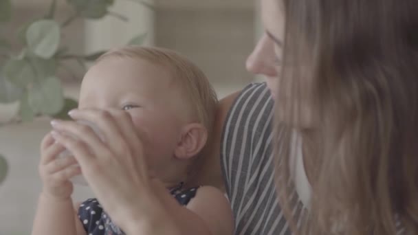 Close-up mooie glimlachende jonge vrouw voeden haar baby meisje uit de kleine baby fles in de keuken. Concept van een gelukkig gezin, één kind, moederschap, liefde. Slow Motion — Stockvideo