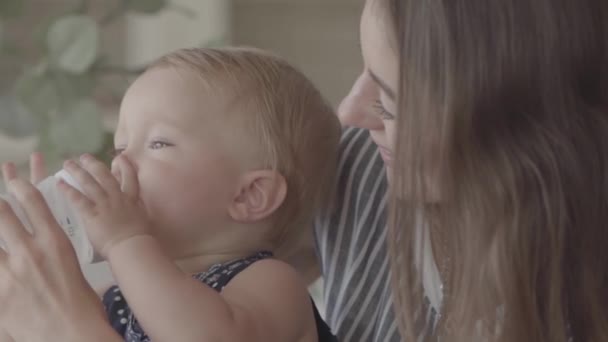 Close-up mooie glimlachende jonge vrouw voeden haar baby meisje uit de kleine baby fles in de keuken. Concept van een gelukkig gezin, één kind, moederschap, liefde. Slow Motion — Stockvideo