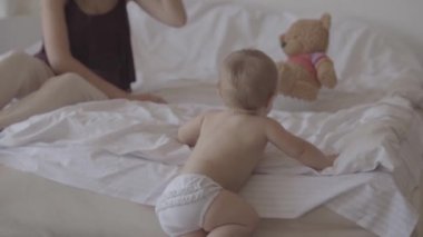 Küçük bebek evde yatakta oynuyor. Bebeğin yanında oturan sevimli anne. Güzel çocuk yatağa tırmanıyor. Mutlu bir aile, bir çocuk, aşk kavramı. Yavaş çekim