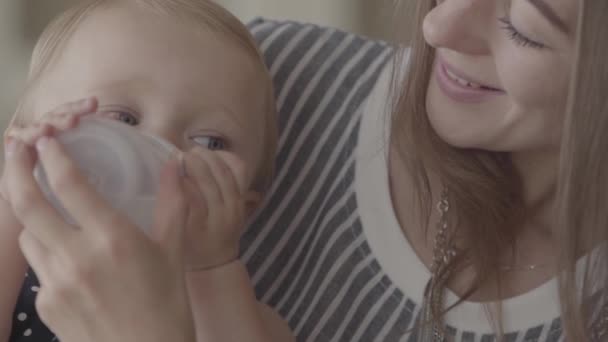 Close-up portret schattig baby meisje drinkwater uit de kleine baby fles met moeders helpen in de keuken. Concept van een gelukkig gezin, één kind, moederschap, liefde. Slow Motion — Stockvideo
