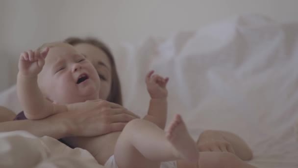 Close-up of a beautiful baby girl crying in mothers arms laying in the bed. A woman trying to calm her naughty child. Concept of motherhood, difficulties in growing kids, love. Slow motion — Stock Video