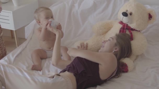 Jonge vrouw zitten met haar baby meisje die spelen met baby fles zittend in het bed, bid Toy Bear is in de buurt. Mooie baby meisje drinkt water. Concept van een gelukkige familie, moederschap. Slow Motion — Stockvideo