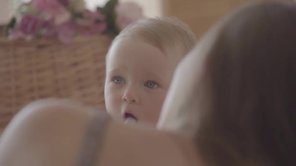 Bonita niña bebe agua del biberón sentada en la cama cerca de mamá en casa. Adorable niño caucásico en el interior — Vídeos de Stock