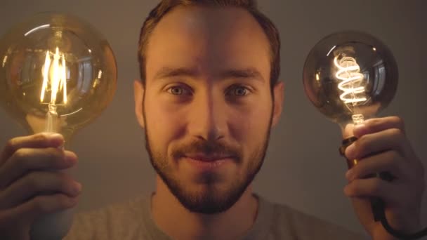 Retrato de cerca del joven barbudo sosteniendo dos bombillas y mirando a la cámara. Concepto de luz y oscuridad, idea, creatividad, electricidad. Serie de personas reales . — Vídeo de stock