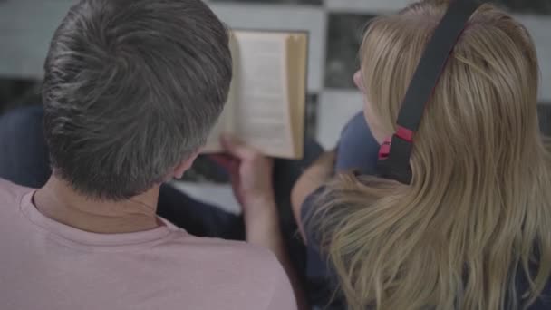 Felnőtt pár időtöltést otthon együtt. A férfi olvasó a könyv és a nő zenét hallgat, akkor változnak. Szabadidő otthon a könyv és a zene szerelmeseinek. Hátulnézet — Stock videók