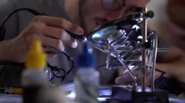 Gözlüklü genç sakallı çırak iş yerinde lehim demiriyle çalışıyor. Adam hata yaptı ve işini düzeltiyor. Meslek kavramı, doğru çalışma, mühendis pozisyonu