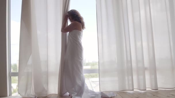 Çekici kadın tabandan tavana pencerenin yanında duran ve uzağa bakan çarşaf kaplı. Kapalı boş zaman, izin, sabah saati — Stok video