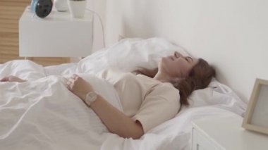 Güzel olgun esmer yavaş yavaş evde sabah uyanma, Uzanır ve yatakta oturur. Güneş büyük pencereden ona parlıyor. Mutlu beyaz kadın yeni günü selamlıyor.
