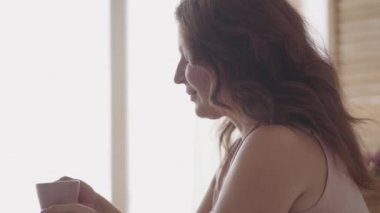 Portre sevimli olgun kadın sabah yatakta oturan çay veya kahve içme. Evde dinlenen gülümseyen bayan. Güneş büyük pencereden ona parlıyor. Mutlu beyaz kadın yeni günü selamlıyor.