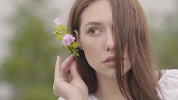 Porträt eines attraktiven jungen brünetten Mädchens mit rosa Blume am Ohr, das wegschaut. Modebegriff, Verbindung zur Natur, ländliches Leben — Stockvideo