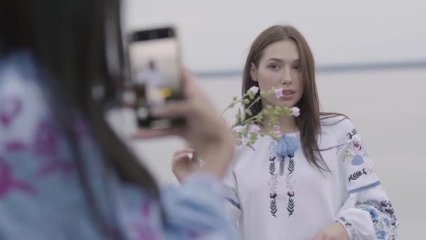 Portret van aantrekkelijk jong brunette meisje in lang geborduurde jurk houden van bloem in de buurt van gezicht poseren terwijl haar vriend foto van haar te nemen. Concept van mode, verbinding met de natuur, landelijk leven — Stockvideo