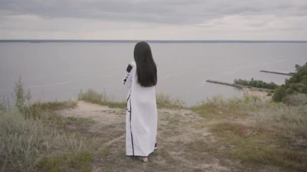 Rückansicht einer entzückenden afrikanisch-amerikanischen Frau in einem langen Sommerkleid, die auf dem Feld vor dem Hintergrund eines Sees oder Flusses spaziert. süße afrikanisch-amerikanische Mädchen verbringen Freizeit im Freien. — Stockvideo