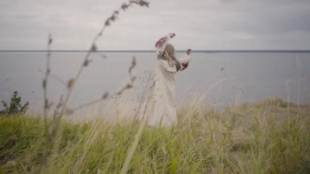 자수 춤과 잔디에 주위를 회전 아름다운 긴 흰색 여름 드레스에 매력적인 금발 소녀. 강이 배경에 있습니다. 패션의 개념, 자연과의 연결 — 비디오