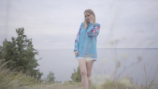 Linda chica caucásica despreocupada que lleva vestido largo de moda de verano que mira con confianza en la cámara disfrutando de los puestos en el fondo del lago. Ocio de una hermosa joven al aire libre. Serie de personas reales . — Vídeos de Stock