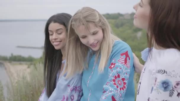 Three girlfriends in beautiful long summer dresses with embroidery sitting and talking on the river bank. Concept of fashion, connection with nature, rural life, friendship — Stock Video