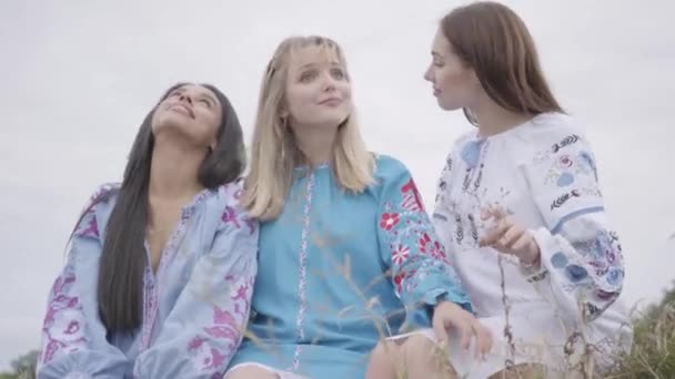 Trois jeunes copines portant une longue robe de mode d'été conversent sur le terrain. Deux filles caucasiennes et une afro-américaine passer du temps libre en plein air . — Video