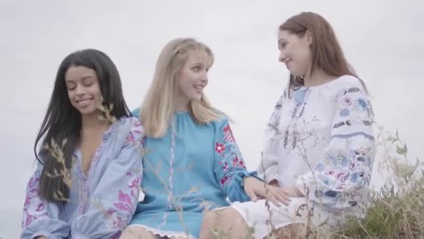 Trois jeunes copines portant une longue robe de mode d'été conversent sur le terrain. Deux filles caucasiennes et une afro-américaine passer du temps libre en plein air . — Video