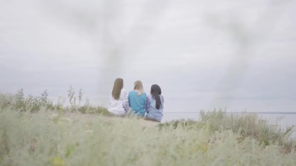 Trois jeunes amies portant une longue robe de mode estivale conversent sur le terrain sur fond de lac ou de rivière. Deux filles caucasiennes et une afro-américaine passer du temps libre en plein air . — Video