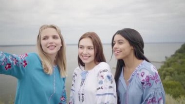 Uzun yaz moda elbise giyen sevimli kız bir göl veya nehir arka planda selfie yapma. İki Kafkas kız ve bir Afrikalı Amerikalı selfie çekerken boş vakit lerini dışarıda geçiriyor