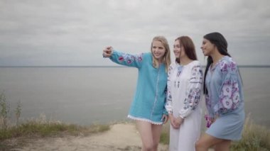 Uzun yaz modası elbisesi giyen üç kız arkadaş bir göl ya da nehrin arka planında selfie çekiyorlar. İki Kafkas kız ve bir Afrikalı Amerikalı selfie çekerken boş vakit lerini dışarıda geçiriyor