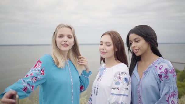 Drie vriendinnen dragen lange zomer mode jurk selfies maken op de achtergrond van een meer of rivier. Twee Kaukasische meisjes en een Afro-Amerikaanse nemen selfie foto besteding vrije tijd buitenshuis — Stockvideo