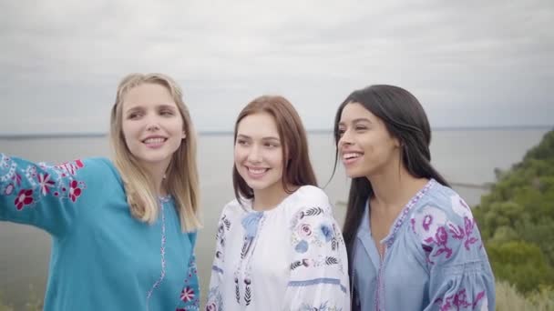 Lindas novias que usan vestido largo de moda de verano haciendo selfies en el fondo de un lago o río. Dos chicas caucásicas y un afroamericano tomando foto selfie pasando tiempo libre al aire libre — Vídeo de stock