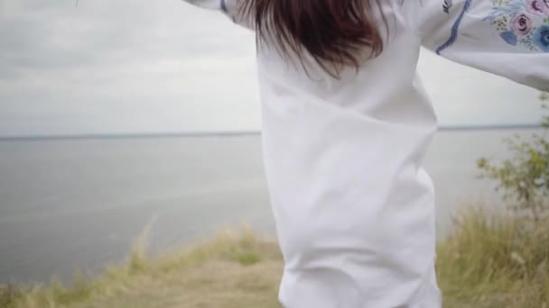 긴 흰색 여름 드레스에 갈색 머리 소녀 잔디의 무리를 들고 높은 잔디 사이에서 실행 자수. 배경에 강. 패션의 개념, 자연과의 연결 — 비디오