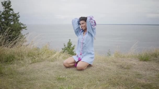 Portrait charmant confiant fille afro-américaine insouciante appréciant assis sur le fond d'un lac ou d'une rivière. Loisirs et week-end d'une jolie jeune femme en plein air. Des vraies séries. Mouvement lent . — Video