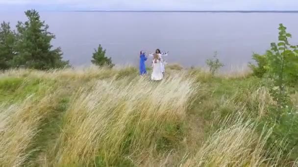 Вид с воздуха на веселые танцующие девушки в длинном летнем платье, прогуливающиеся вдоль озера или реки. Две белые и одна афроамериканка проводят досуг на свежем воздухе. Стрельба дронами . — стоковое видео