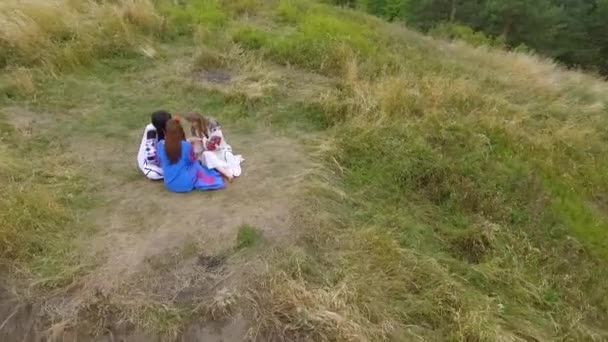 Nehirdeki yüksek tepede çimenlerde oturan nakışlı uzun elbiseleriçinde üç kız arkadaşın havadan görünümü. Doğayla bağlantı, kırsal yaşam, dostluk. Drone çekim, üst tegörüş — Stok video