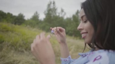 Yakın çekim portre büyüleyici kaygısız Afro-Amerikan kız uzun yaz moda elbise giyen alanında duruyor zevk. Açık havada çiçek açan bir kır çiçeği tutan güzel bir genç kadının eğlencesi.