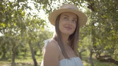 Çekici genç kadın hasır şapka lı ve uzun beyaz elbiseli yeşil yaz bahçesinde gülümseyerek kameraya bakıyor. Kamera uzaklaşıyor