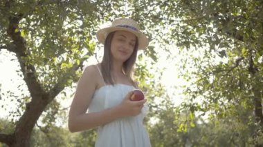 Çekici genç kadın hasır şapka ve uzun beyaz elbise yeşil yaz bahçesinde ayakta elma tutan kameraya bakıyor. Hasat zamanı, kırsal yaşam tarzı