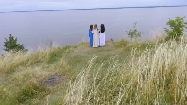 Vista aérea a tres novias que usan vestido largo de la manera del verano que camina cerca del lago o del río. Dos caucásicas y una afroamericana pasan tiempo libre al aire libre. Disparos con drones . — Vídeo de stock