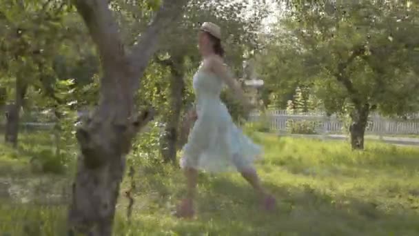 Jolie jeune femme en chapeau de paille et longue robe blanche traversant le jardin d'été vert. Vie rurale sans soucis, connexion avec la nature. Caméra en mouvement près 