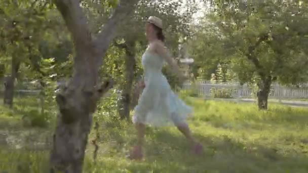 Atractiva joven con sombrero de paja y vestido blanco largo corriendo por el verde jardín de verano. Vida rural despreocupada, conexión con la naturaleza. Cámara moviéndose cerca — Vídeos de Stock