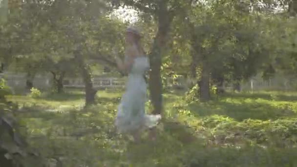 Carina giovane donna in cappello di paglia e lungo abito bianco che corre attraverso il verde giardino estivo. Vita rurale spensierata, connessione con la natura . — Video Stock