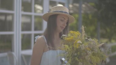 Hasır şapkalı ve beyaz elbiseli çekici genç kadın küçük köy evinin önünde kır çiçekleri koklarken kameraya gülümseyerek bakıyor. Kırsal yaşam tarzı