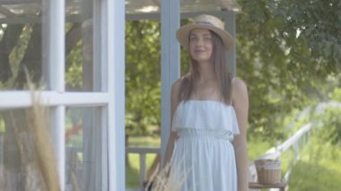 Hasır şapkalı ve uzun beyaz elbiseli çekici genç, küçük köy evinin önünden bakan genç kadın. Kaygısız kırsal yaşam, doğa ile bağlantı