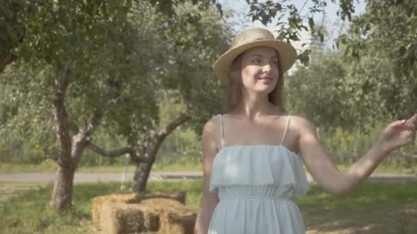 Linda joven sonriente mujer en sombrero de paja y vestido blanco largo caminando a través del verde jardín de verano. Vida rural despreocupada, conexión con la naturaleza. Vista frontal — Vídeos de Stock