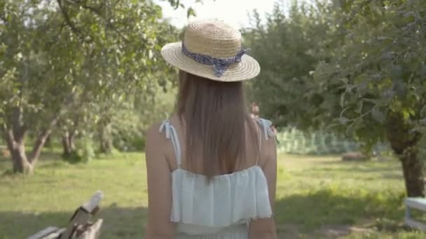 Vista posterior de la linda mujer joven en sombrero de paja y vestido blanco largo caminando a través del verde jardín de verano. Vida rural despreocupada, conexión con la naturaleza . — Vídeos de Stock