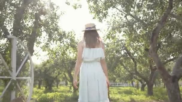Saman şapkalı ve yeşil yaz bahçesinde yürüyen uzun beyaz elbiseli güzel genç bir kadının arka görünümü. Kaygısız kırsal yaşam, doğa ile bağlantı. — Stok video