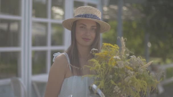 Adorabile giovane donna in cappello di paglia e vestito bianco guardando la fotocamera sorridente mentre annusa fiori selvatici di fronte alla piccola casa di paese. Stile di vita rurale — Video Stock