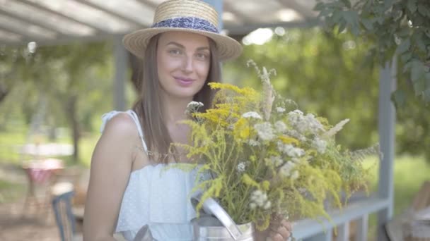 Ritratto bella giovane donna in cappello di paglia e vestito bianco guardando la macchina fotografica sorridente mentre tiene fiori selvatici. Stile di vita rurale . — Video Stock