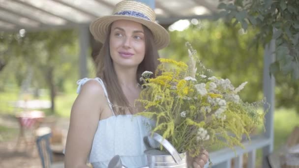 Retrato hermosa mujer joven en sombrero de paja y vestido blanco mirando a la cámara sonriendo mientras sostiene flores silvestres. Estilo de vida rural . — Vídeos de Stock