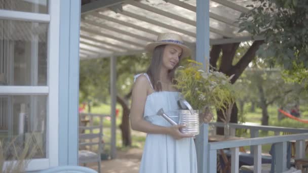 Jolie jeune femme en chapeau de paille et robe blanche souriante tout en reniflant des fleurs sauvages dans un arrosoir en face de la petite maison de village. Mode de vie rural — Video