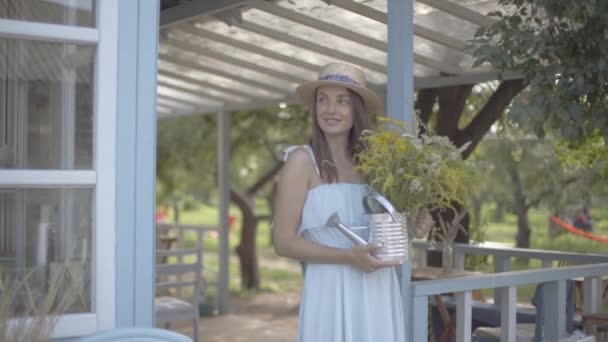 Mulher jovem bonito em chapéu de palha e vestido branco sorrindo enquanto farejando flores silvestres em uma lata de rega na frente da pequena casa da aldeia. Estilo de vida rural — Vídeo de Stock