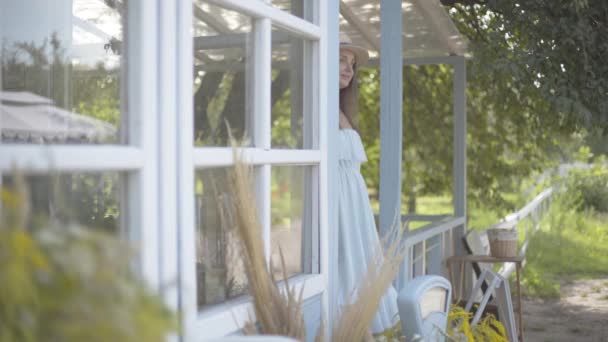 Adorable young smiling woman in straw hat and long white dress looking away in front of the small village house. Carefree rural life, connection with nature — Stock Video